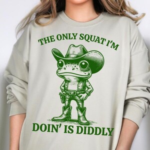 Może przedstawiać: Jasnozielona bluza z napisem "The only squat I'm doin' is diddly" i kresk&oacute;wkową żabą w kowbojskim kapeluszu i butach.