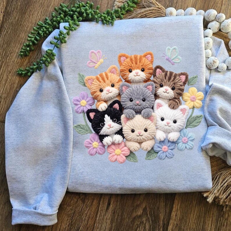 Pu&ograve; includere: Felpa azzurra con un ricamo di sette adorabili gattini in vari colori, circondati da fiori e farfalle. I gattini includono gatti arancioni, grigi, neri e bianchi e tabby marroni. Il disegno &egrave; centrato sul davanti.