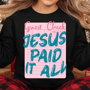 May include: Black sweatshirt featuring a pink guest check design with the words "Guest Check" and "JESUS PAID IT ALL" in teal. The design includes a check number. A cozy and inspirational apparel item.