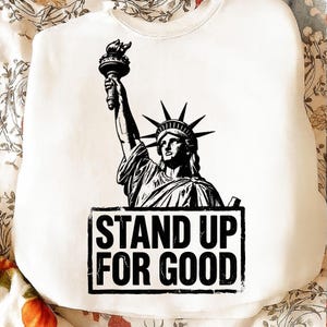 May include: White sweatshirt featuring a black and white graphic of the Statue of Liberty holding a torch. The text "STAND UP FOR GOOD" is printed in a black rectangle below the statue. The sweatshirt is made of a soft material.