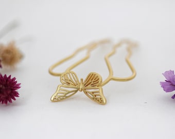 Puede incluir: Un peine para el cabello de color dorado con un diseño de mariposa. La mariposa tiene intrincados detalles recortados. El peine está sobre un fondo blanco, con flores moradas y marrones a ambos lados.