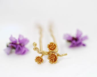 Puede incluir: Una horquilla dorada con un diseño floral, con tres racimos de flores en una rama. La horquilla se encuentra sobre un fondo blanco, con flores moradas borrosas a ambos lados.