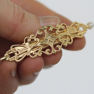 Könnte beinhalten: Eine goldfarbene Haarklammer mit einem zarten, verzierten Design. Die Klammer hat ein zentrales, geometrisches Muster und eine kleine Perle als Akzent am Ende.