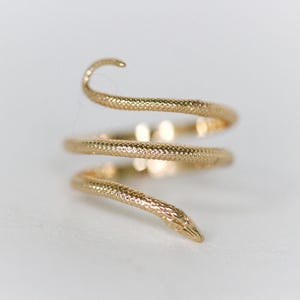 May include: A gold-colored snake ring with a textured, scale-like pattern. The ring coils around the finger in a spiral, with the snake's head and tail forming the ends. The ring is set against a white background.