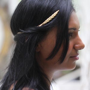 Puede incluir: El cabello oscuro de una mujer está adornado con una horquilla dorada en forma de espiga de trigo. El cabello está peinado con una raya a un lado, y algunos mechones enmarcan la cara. El fondo está borroso.