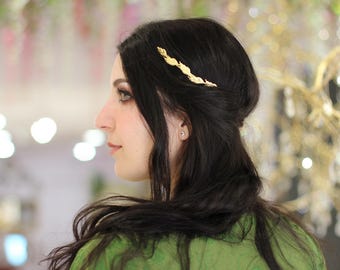 Puede incluir: Un accesorio para el cabello en tono dorado con un diseño de rama delgada y alargada con detalles texturizados en forma de hojas. La pieza metálica se coloca horizontalmente en el lateral del cabello oscuro para sujetar un mechón.