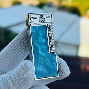 May include: A silver lighter with a rectangular shape and a vibrant blue marbled design. The top of the lighter has the word "PARIS" and "1898" printed on it. The lighter is held in a white glove.