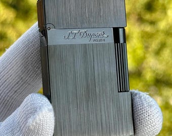 May include: A dark gray S.T. Dupont lighter with a textured, brushed metal finish. The lighter features the brand name "S.T. Dupont Paris" in a silver tone. The design includes a vertical ribbed section on the side.