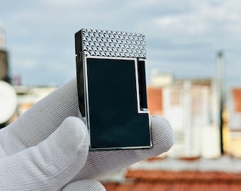 May include: A rectangular, silver and black lighter with a geometric pattern on the top. The lighter has a sleek, modern design and is held by a gloved hand against a blurred outdoor background.