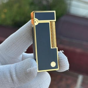 May include: A black and gold lighter with a rectangular design. The lighter features a gold frame and accents, with a black central panel. It has a textured, gold-coloured ignition wheel and a small, circular emblem on the front.