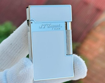 May include: A silver and white S.T. Dupont lighter. The lighter has a rectangular shape with rounded edges and a hinged top. The brand name "S.T. Dupont PARIS" is engraved on the front. The lighter is held by a gloved hand.