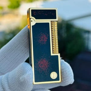 May include: A gold and black rectangular lighter. The lighter features a gold frame, a black body with red speckles, and a gold cylindrical detail. It has a small circular emblem at the base. The design is elegant and compact.