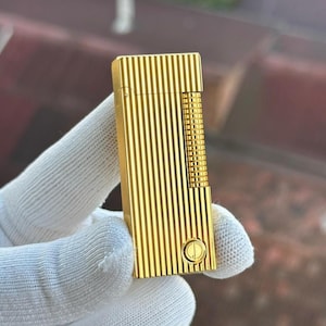 May include: A gold-coloured lighter with vertical lines and a textured rectangular section. The lighter has a round button on the bottom and is held by a gloved hand. The lighter is a luxury item.