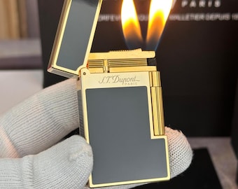 Könnte beinhalten: Ein goldfarbenes Feuerzeug mit einem schwarzen rechteckigen Feld und dem Markennamen "S.T. Dupont" wird mit seiner Flamme gezeigt. Das Feuerzeug wird von einer behandschuhten Hand gehalten, und der Hintergrund ist dunkel.
