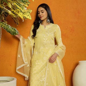 May include: A yellow embroidered kurta with a matching dupatta and dhoti pants. The kurta has a V-neckline and is decorated with white floral embroidery. The dupatta is sheer and has a lace trim. The dhoti pants are also yellow and have a lace trim.