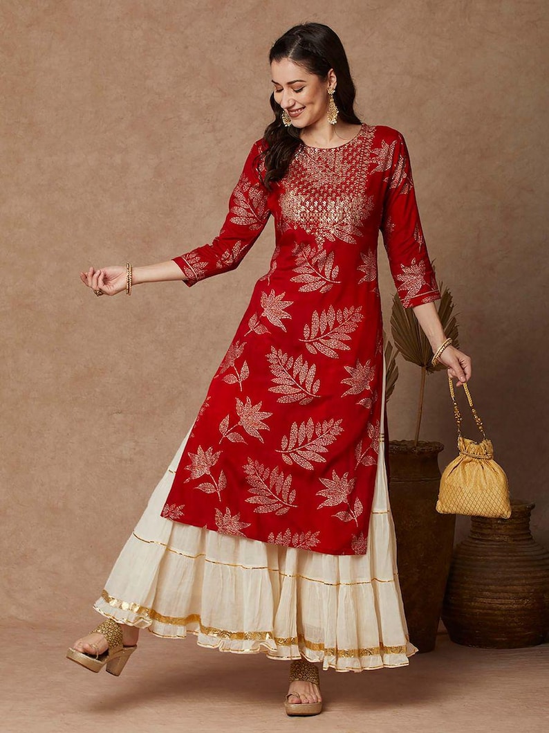 May include: A red and gold kurta with a leaf print and a white layered skirt. The kurta has a round neckline and three-quarter sleeves. The skirt has a gold trim.