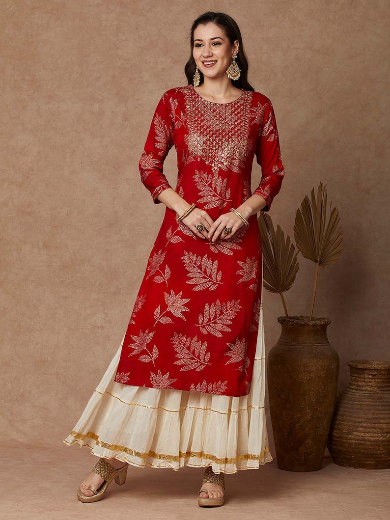 May include: A red and gold kurta with a leaf print and a white layered skirt. The kurta has a round neckline and three-quarter sleeves. The skirt has a gold trim.