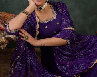 Puede incluir: Una mujer con un conjunto salwar kameez morado. El atuendo presenta un dupatta estampado y ribetes dorados. La parte superior tiene un escote cuadrado con bordados dorados. Lleva un collar de perlas y pulseras.