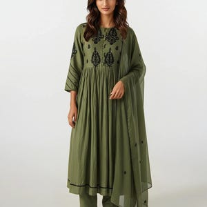 May include: An olive green ensemble featuring a long-sleeved dress with black embroidered details on the bodice and sleeves, paired with matching pants and a sheer scarf. The outfit is completed with white sneakers. The dress has a flowing, pleated skirt.