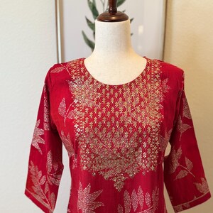 May include: A red kurta with a gold embroidered neckline and leaf pattern. The kurta is made of a soft, lightweight fabric and has a relaxed fit.
