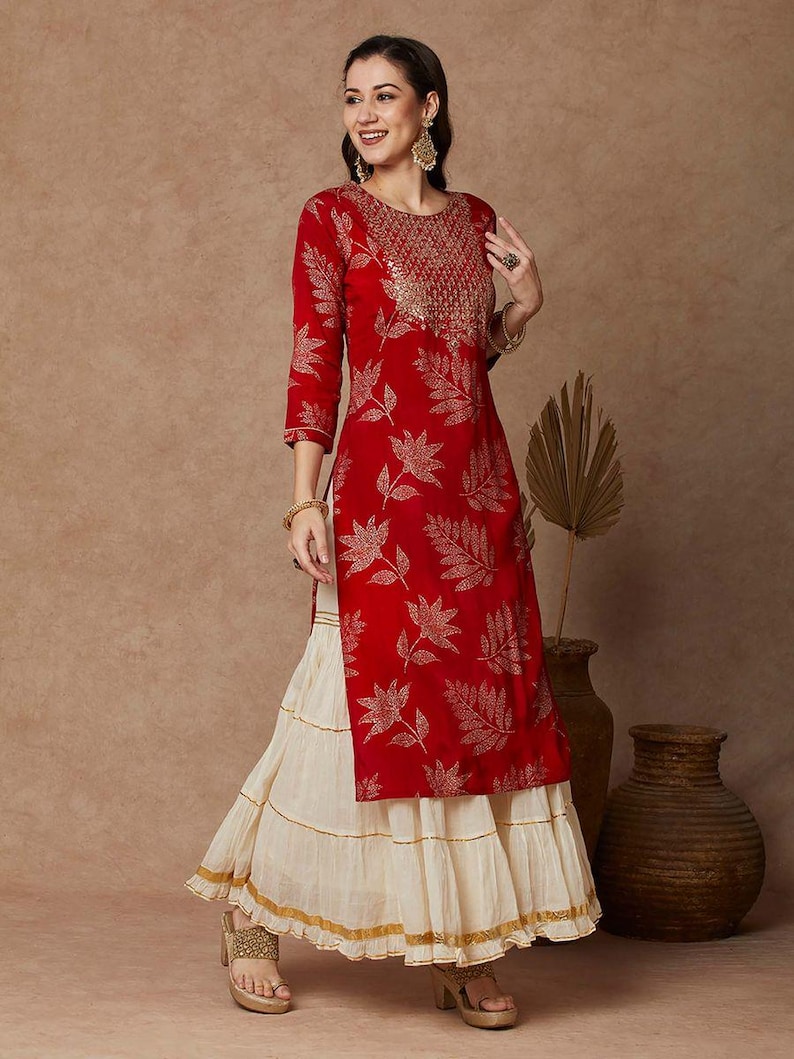 May include: A red long-sleeved kurta with a gold leaf print and gold embroidery around the neckline. The kurta is paired with a white layered skirt with gold trim.