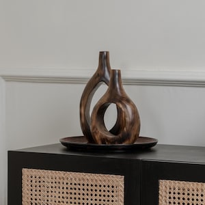 Può includere: Due vasi decorativi in legno dalle forme organiche poggiano su un piatto marrone scuro. I vasi sono posti sopra un mobile nero con pannelli porta in canna intrecciata. Una foglia è visibile a destra.