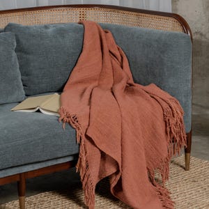 Op de afbeelding: Een terracotta geweven sprei met franjes is over een blauwgroene bank gedrapeerd. Een open boek ligt op de bank. De bank heeft een houten frame en een geweven rugleuning. Het vloerkleed is gemaakt van geweven natuurlijke vezels.