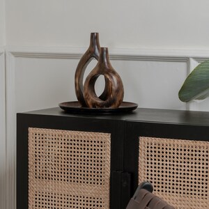 May include: Two wooden decorative vases with organic shapes sit on a dark brown plate. The vases are placed on top of a black cabinet with woven cane door panels. A leaf is visible on the right side of the image.