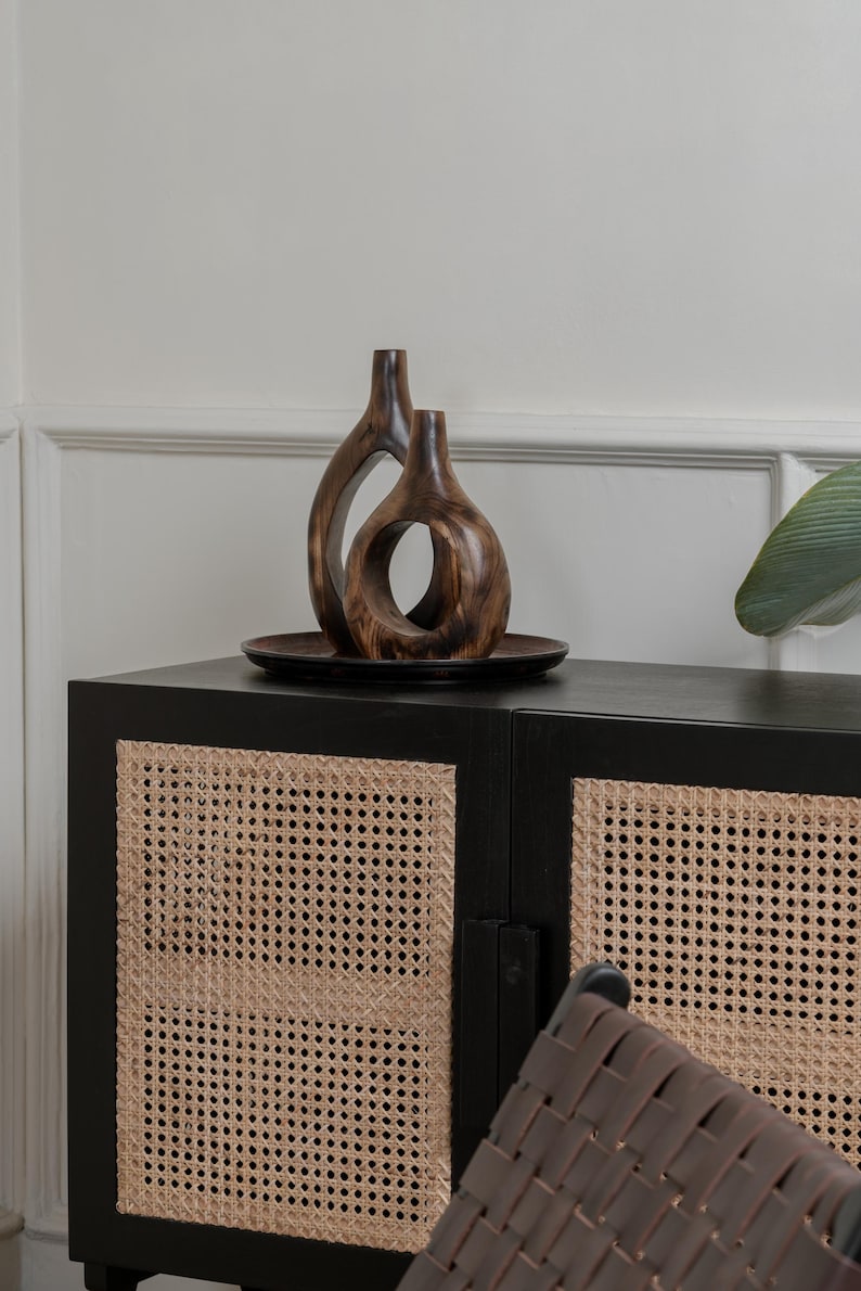 May include: Two wooden decorative vases with organic shapes sit on a dark brown plate. The vases are placed on top of a black cabinet with woven cane door panels. A leaf is visible on the right side of the image.