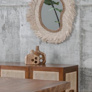 May include: A decorative mirror with a natural fiber frame and fringe hangs above a wooden cabinet. Two wooden vases sit on the cabinet. A wooden table and chairs are in the foreground. The room has a modern, minimalist aesthetic.