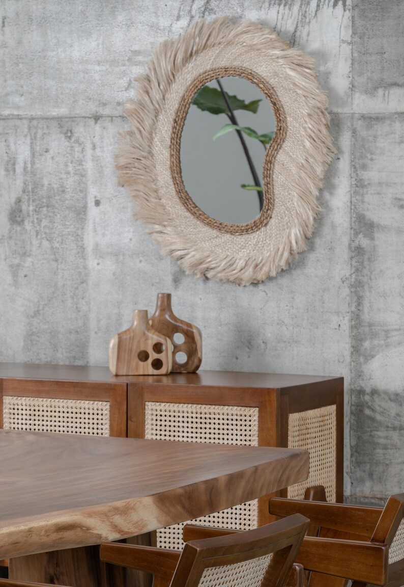May include: A decorative mirror with a natural fiber frame and fringe hangs above a wooden cabinet. Two wooden vases sit on the cabinet. A wooden table and chairs are in the foreground. The room has a modern, minimalist aesthetic.
