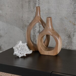 May include: Two wooden vases with circular cutouts and a white seashell sit on a black surface. The vases are light brown and have a smooth finish. The seashell is intricately detailed.