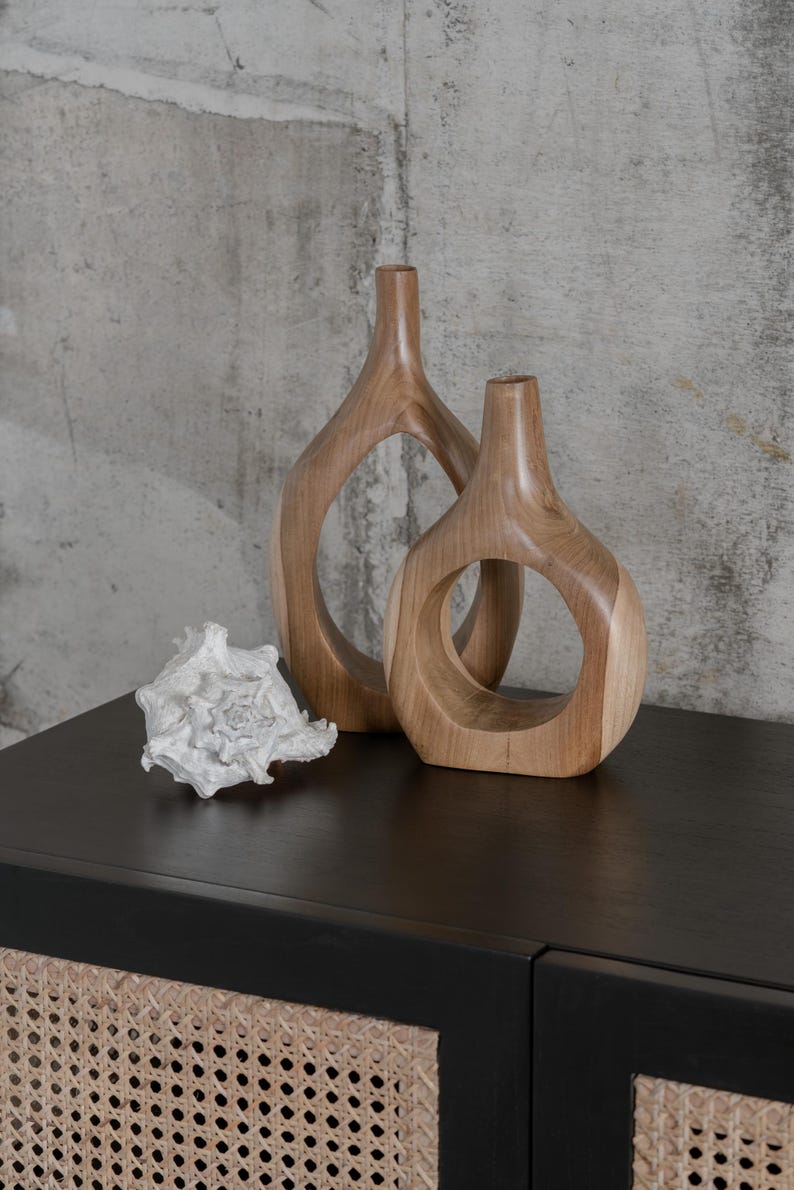 May include: Two wooden vases with circular cutouts and a white seashell sit on a black surface. The vases are light brown and have a smooth finish. The seashell is intricately detailed.