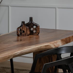 May include: Two dark brown wooden decorative vases with circular cutouts sit on a large wooden table. The table has a natural edge and is supported by black metal legs. Black chairs are partially visible.