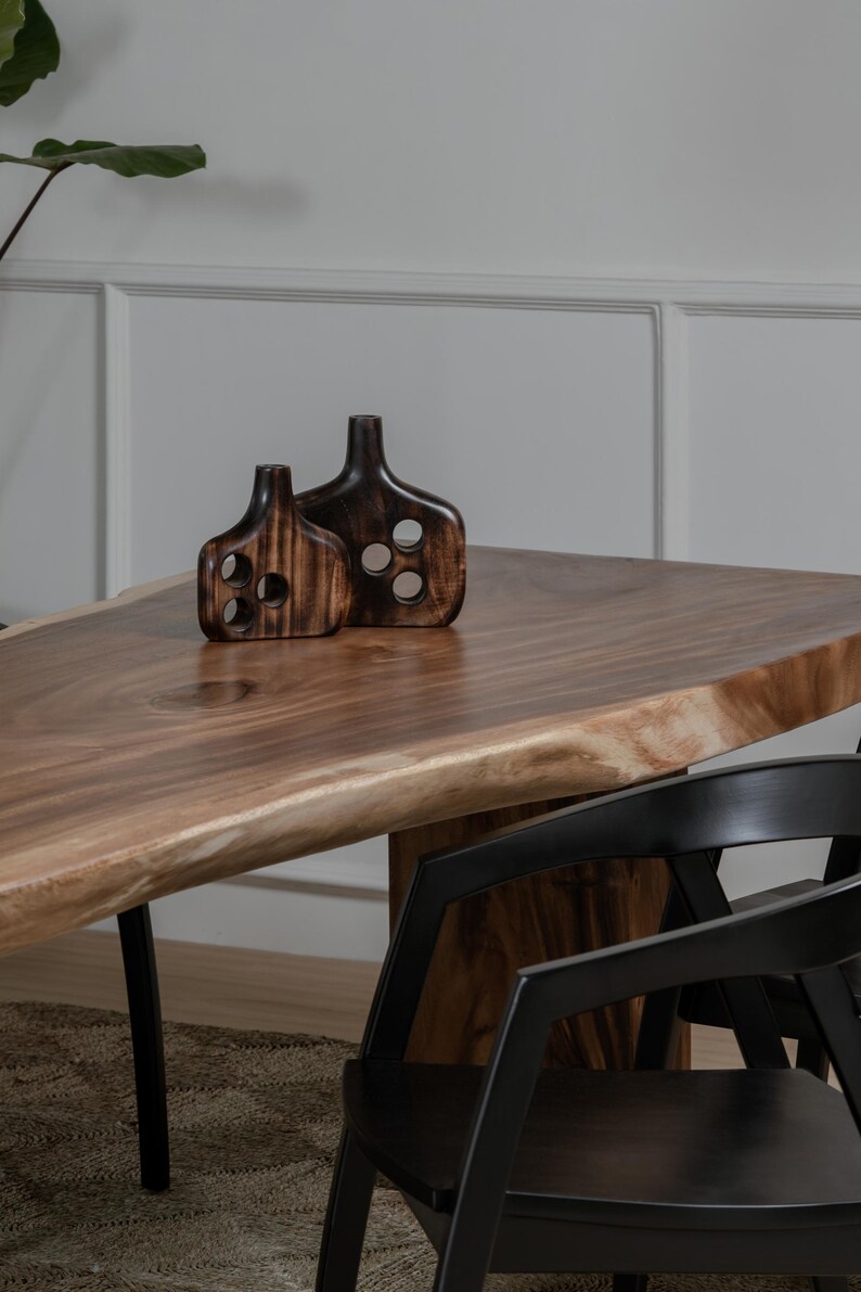 May include: Two dark brown wooden decorative vases with circular cutouts sit on a large wooden table. The table has a natural edge and is supported by black metal legs. Black chairs are partially visible.