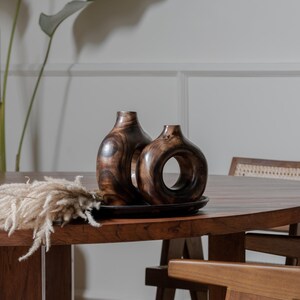 May include: Two wooden decorative vases with a dark brown finish, one with a circular opening, sit on a dark tray. The vases are on a wooden table with a dried floral arrangement. The table and chairs are made of wood.