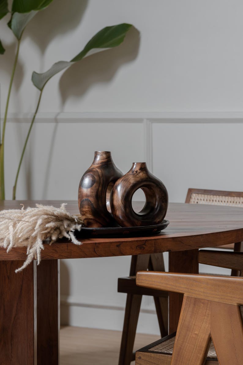 May include: Two wooden decorative vases with a dark brown finish, one with a circular opening, sit on a dark tray. The vases are on a wooden table with a dried floral arrangement. The table and chairs are made of wood.