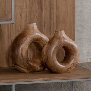 May include: Two wooden vases with a distinctive circular design. The vases are crafted from light brown wood and have a smooth, polished finish. They are displayed on a wooden shelf against a wooden panel background.