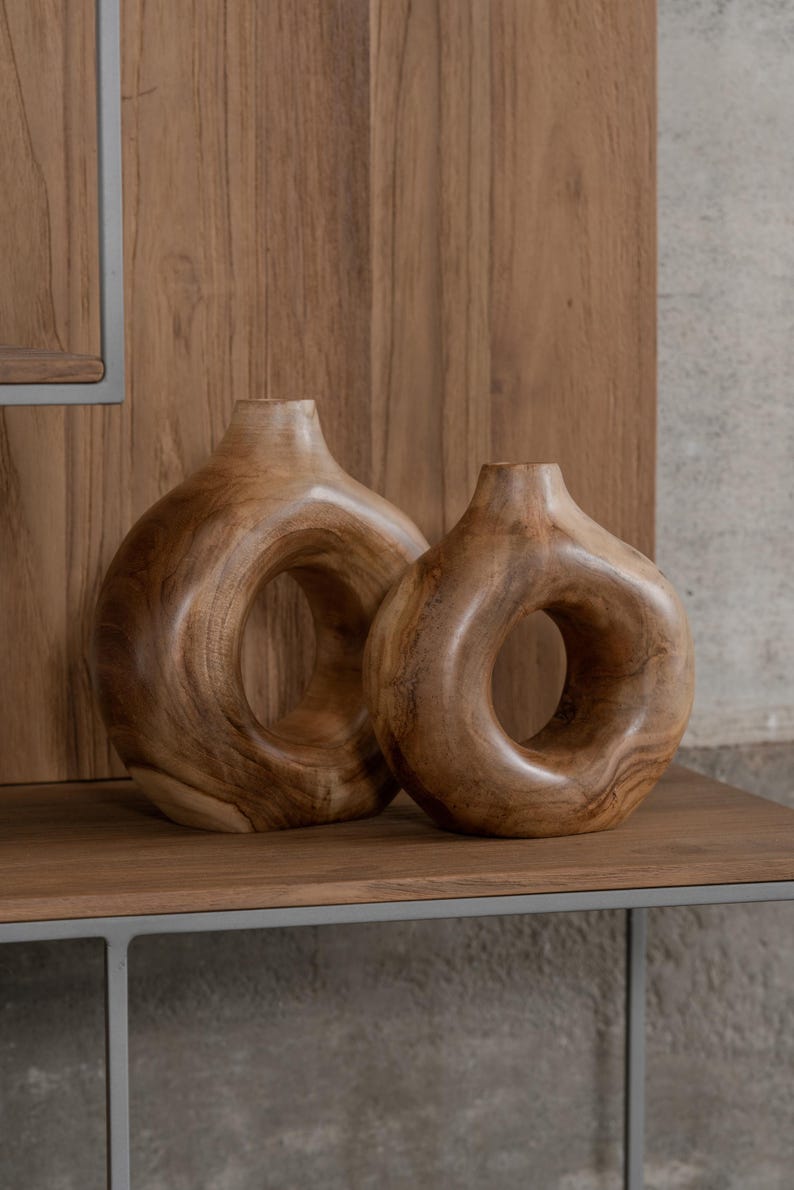 May include: Two wooden vases with a unique circular design. The vases are made of a light brown wood and have a smooth, polished finish. They are displayed on a wooden shelf against a wooden panel background.
