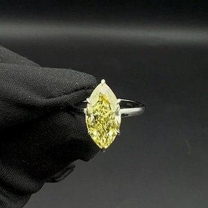 May include: A marquise-cut yellow diamond ring. The diamond is set on a thin, silver band. The ring is held against a black background. The diamond has a bright, vibrant colour and a pointed shape.