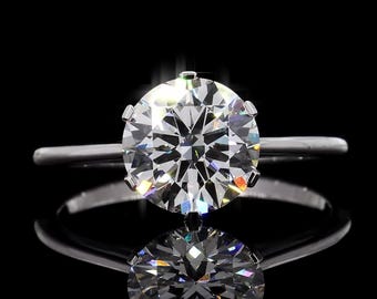 May include: A solitaire engagement ring with a large, round-cut diamond set in a simple, silver band. The diamond sparkles brightly against a black background, with its reflection visible below. The ring is a classic symbol of love and commitment.