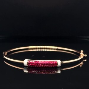 May include: A gold bangle bracelet featuring a rectangular section of red gemstones set in a white enamel frame. The bracelet is shown on a reflective black surface.