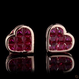 May include: A pair of heart-shaped stud earrings. Each earring has a gold-coloured frame filled with square-cut, deep red gemstones. The earrings are set against a black background, with their reflections visible below.