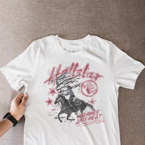 Pode incluir: Camiseta branca com um gráfico desgastado que apresenta um cowboy montando um cavalo, uma bandeira americana e os dizeres "Hellstar" e "Too Wild Just Heat" em vermelho. A camiseta tem mangas curtas.