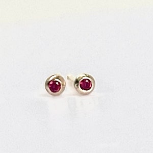 May include: A pair of small stud earrings. Each earring features a round, deep red gemstone set in a silver bezel. The earrings are displayed on a white surface, showcasing their simple elegance.
