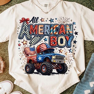 Op de afbeelding: Een crèmekleurig T-shirt met korte mouwen en een grafische print met de tekst All American Boy. Het ontwerp bevat een blauwe monstertruck met een rood-wit-blauwe vlag, sterren, vuurwerk en een rode strik in een patriottisch kleurenschema.