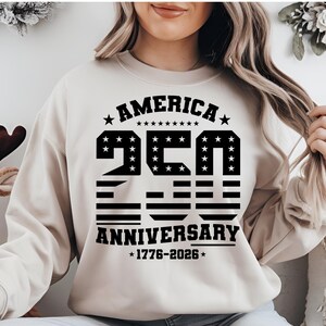 Op de afbeelding: Een beige sweater met ronde hals en een zwarte grafische print met de tekst AMERICA 250 ANNIVERSARY 1776-2026. Het ontwerp bevat sterren en horizontale strepen in de grote cijfers 250, omlijst door tekst en decoratieve sterren.