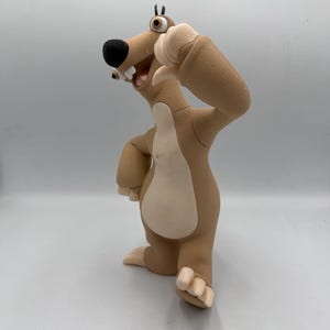 May include: A tan and cream-coloured sloth figurine with a large black nose, wide-set eyes, and three white teeth. The character has a textured surface, stands on two feet with visible claws, and holds one arm raised with a thumb pointing downward.