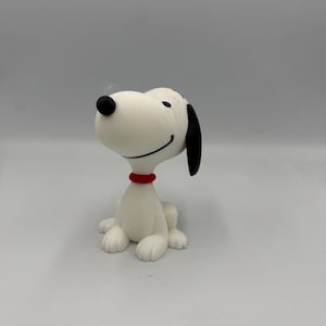 May include: A white dog figurine with a black nose, black ears, and a red collar. The dog is in a sitting position with a smiling expression, featuring smooth, matte surfaces and a minimalist design that captures the iconic cartoon character.