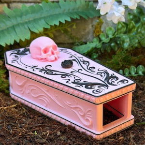 Può includere: Una scatola rosa chiaro a forma di bara con un teschio rosa in rilievo e una piccola rosa nera sul coperchio. Il coperchio è decorato con motivi in filigrana neri e i lati della scatola presentano dettagli a volute in rilievo.